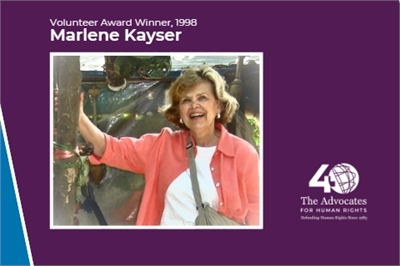 [ai] A woman with curly hair smiling and standing in front of a colorful backdrop. Text on the image identifies her as Marlene Kayser, Volunteer Award Winner in 1998, accompanied by a logo for The Advocates for Human Rights.